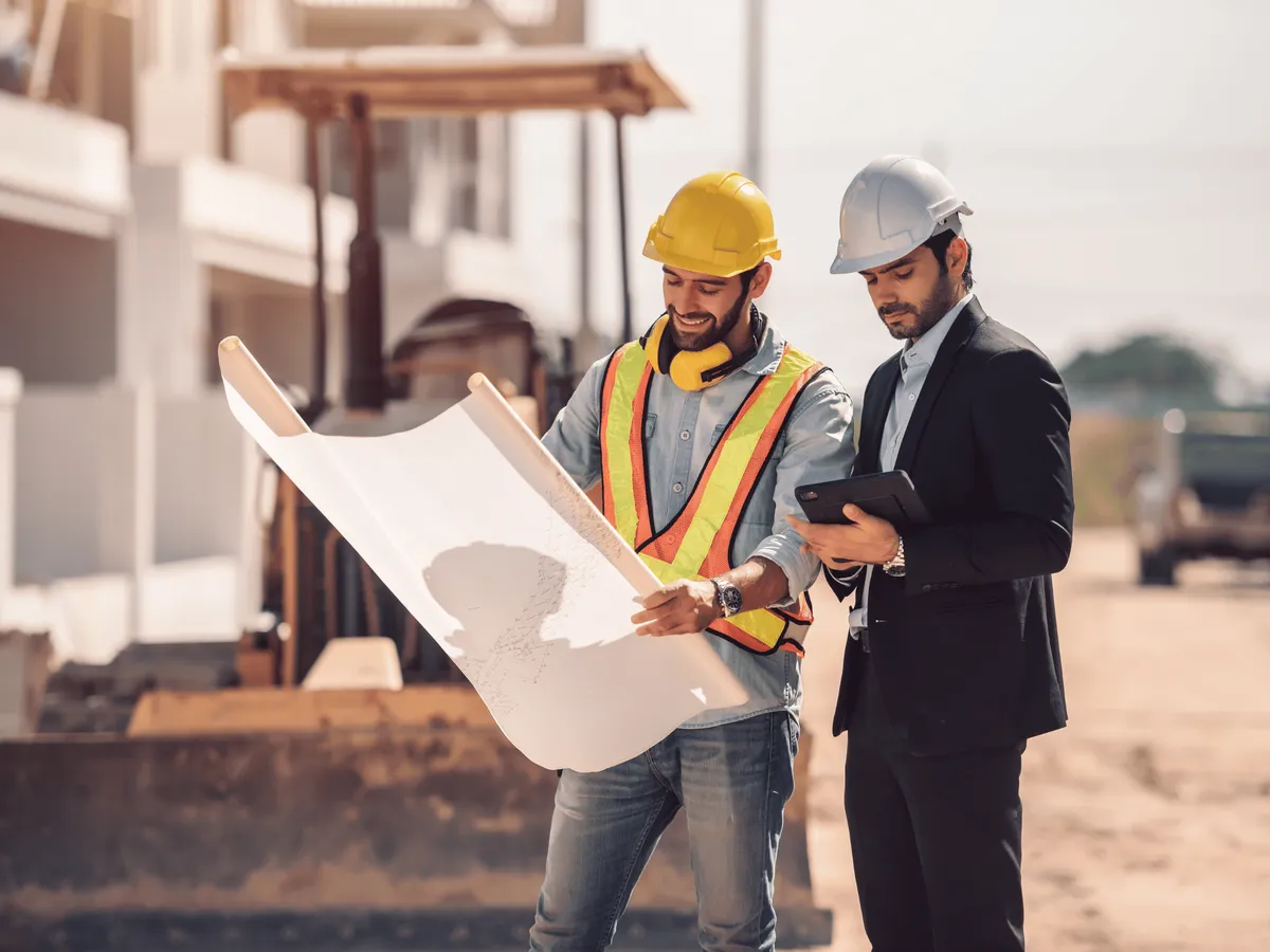 Bauleiter und Logistiker mit gelbem und weißem Helm besprechen Pläne auf einer großen Baustelle vor Seecontainern